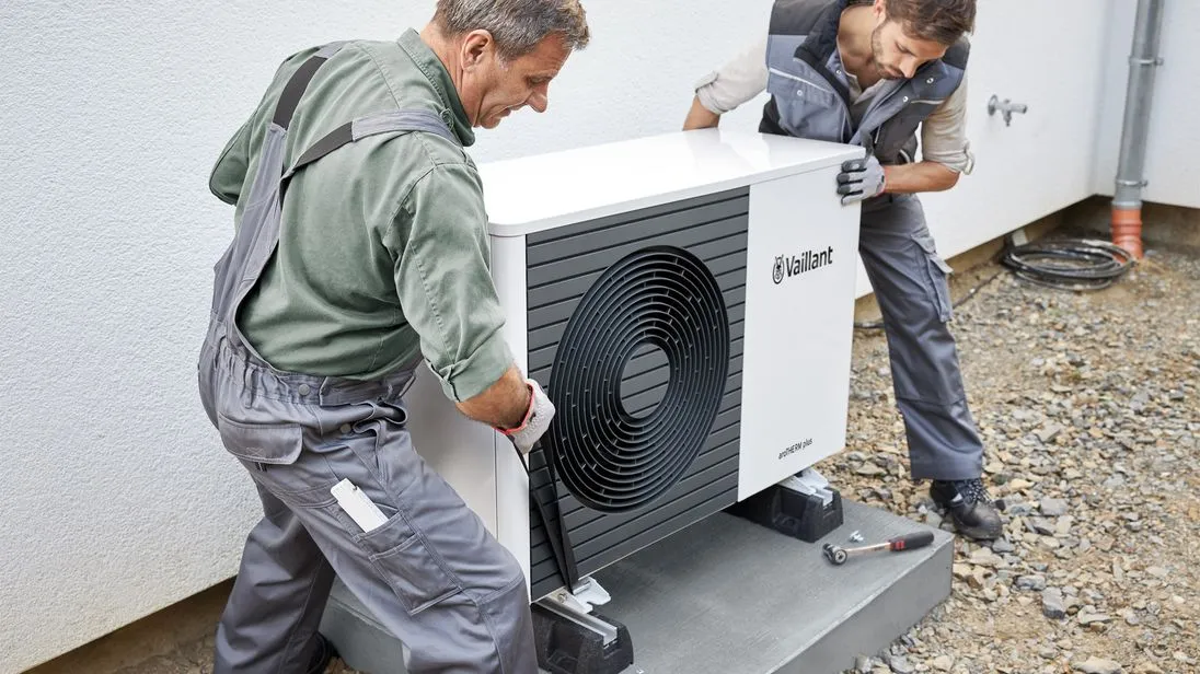 Zwei Techniker in Arbeitskleidung installieren eine Wärmepumpe vor einem Wohnhaus.