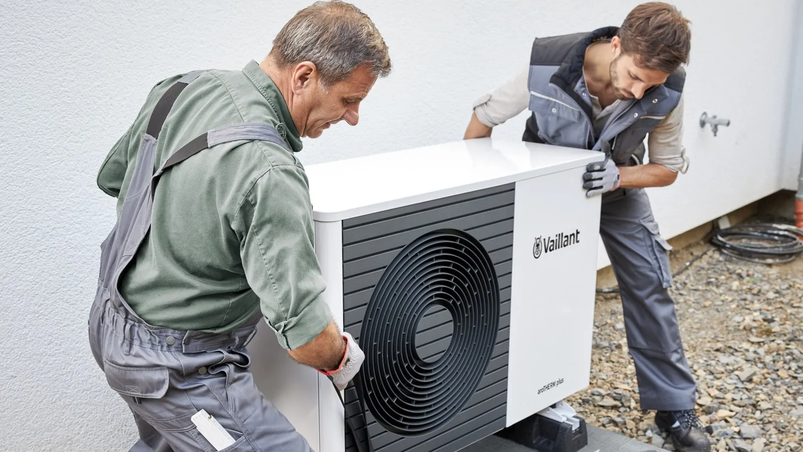 Zwei Techniker in Arbeitskleidung installieren eine Wärmepumpe vor einem Wohnhaus.