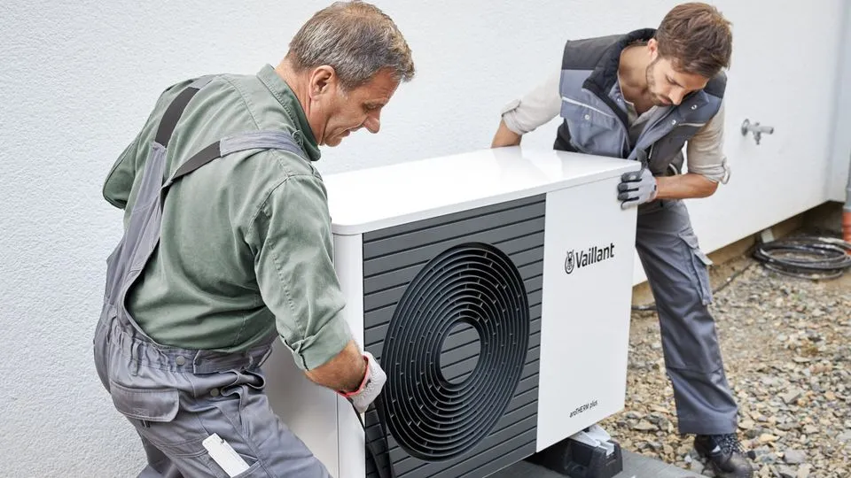 Zwei Techniker in Arbeitskleidung installieren eine Wärmepumpe vor einem Wohnhaus.