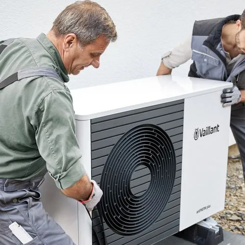 Zwei Techniker in Arbeitskleidung installieren eine Wärmepumpe vor einem Wohnhaus.