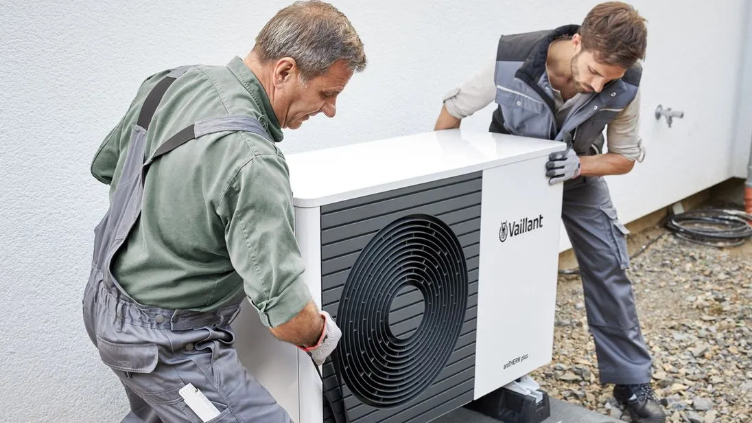 Zwei Techniker in Arbeitskleidung installieren eine Wärmepumpe vor einem Wohnhaus.