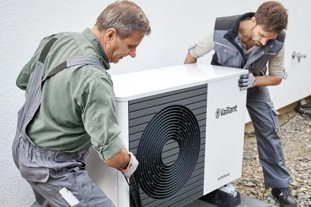 Zwei Techniker in Arbeitskleidung installieren eine Wärmepumpe vor einem Wohnhaus.